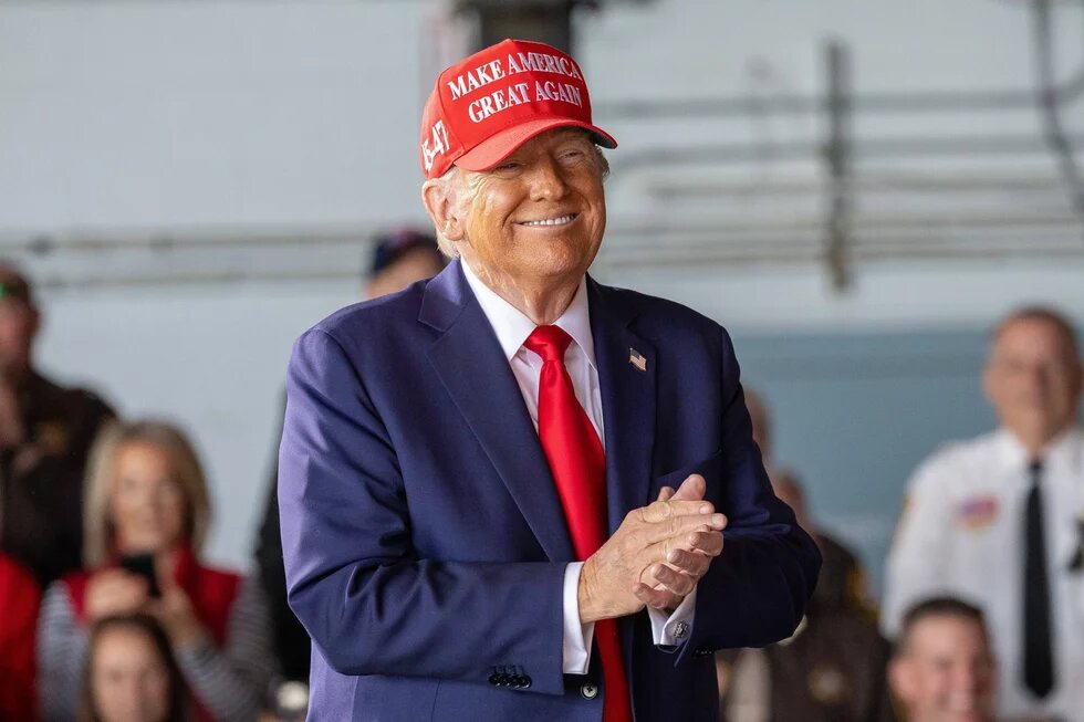 Foto: Donald Trump mit roter „Make America Great Again“-Kappe und Anzug reibt sich grinsend die Hände. Unscharfes Publikum im Hintergrund.