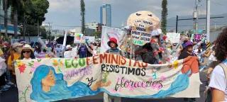 Mujeres marchando con una manta en la que se leé "Ecofeministas, juntas resistimos y florecemos"
